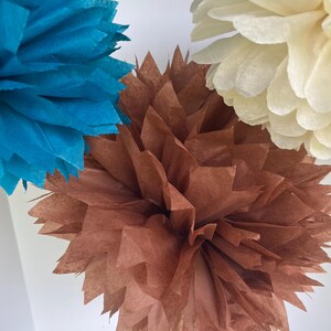 May include: Three decorative paper pom-poms in teal, brown, and cream. Each pom-pom is constructed from multiple layers of thin, folded tissue paper with pointed, jagged edges, creating a full, spherical, and textured floral-like appearance.