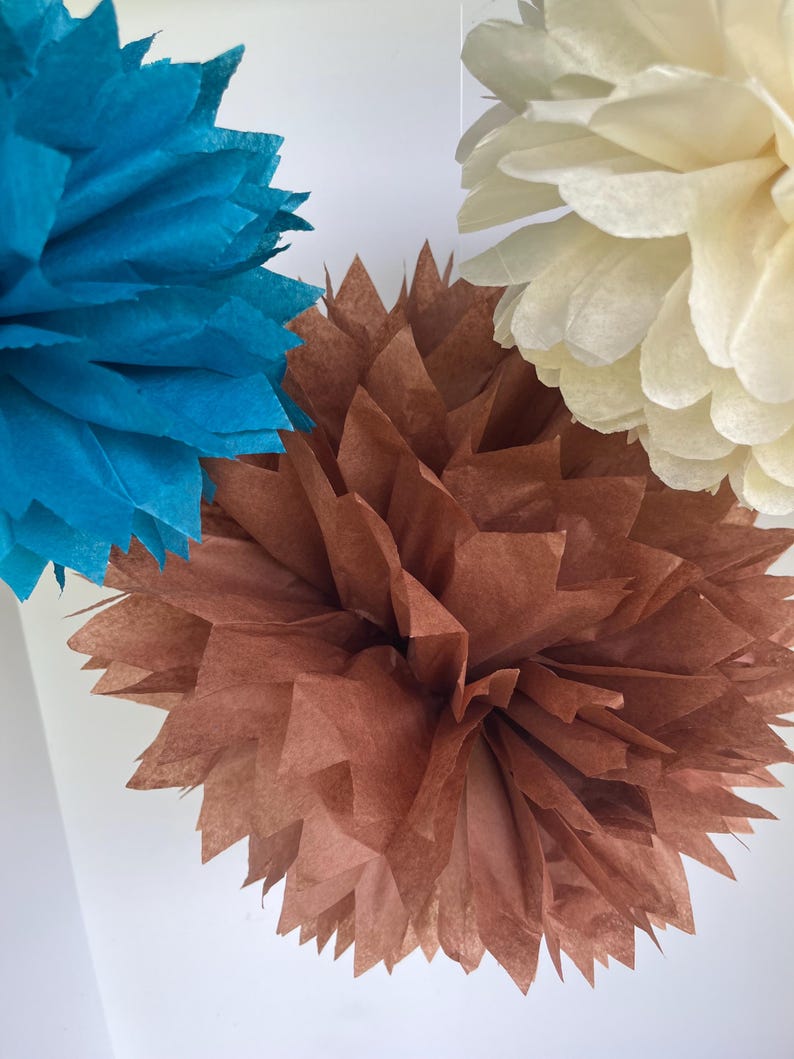 May include: Three decorative paper pom-poms in teal, brown, and cream. Each pom-pom is constructed from multiple layers of thin, folded tissue paper with pointed, jagged edges, creating a full, spherical, and textured floral-like appearance.