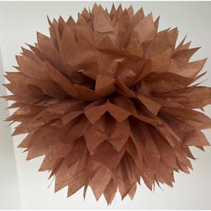 May include: A spherical paper pom-pom decoration in a solid brown color. The object is constructed from multiple layers of thin, translucent paper sheets with pointed, jagged edges that fan out to create a full, textured, and voluminous floral shape.