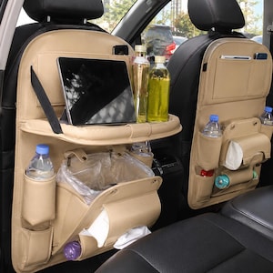 May include: Two beige car seat organizers attached to the back of front vehicle seats. Each organizer features multiple storage pockets, a fold-down tray table holding a tablet, bottle holders, and a compartment containing a plastic trash bag.