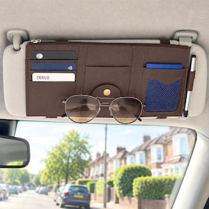 May include: A brown car visor organizer featuring multiple card slots, a blue mesh pocket, a pen holder, and a central snap-button strap for sunglasses. The organizer is attached to a light gray vehicle sun visor and contains several cards.
