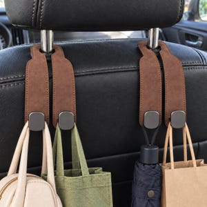 May include: Two brown faux leather car seat headrest hooks with dark metallic hardware. Each hook features a dual-strap design that loops around the headrest posts, providing four individual hooks to hang bags, an umbrella, and other personal items.