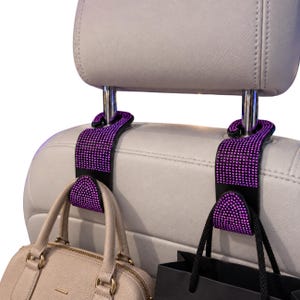 May include: Two black car headrest hooks covered in small, sparkling purple rhinestones. Each hook is attached to the metal posts of a light gray car headrest. A beige handbag hangs from the left hook, and a black shopping bag hangs from the right.