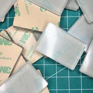 Könnte beinhalten: Eine Ansammlung rechteckiger, durchscheinender Kunststoffplättchen mit beigefarbener Kleberückseite. Jedes Teil ist auf der Rückseite mit einem 3M 468MP 200MP Klebeetikett versehen. Die Plättchen liegen überlappend verstreut und zeigen sowohl die glatten, klaren Vorderseiten als auch die mit beigem Papier bedeckten Klebeseiten.