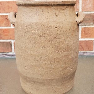 May include: A cylindrical terracotta vessel with a rough, textured surface and a wide opening. The vessel features two small, curved handles positioned on opposite sides near the top rim, showcasing a natural, earthy brown clay finish throughout.