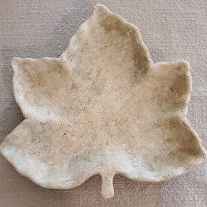 May include: A leaf-shaped dish with a textured, matte surface in shades of beige, cream, and light brown. The object features irregular, raised edges and a distinct stem at the base, mimicking the organic form of a dried maple leaf in a neutral tone.