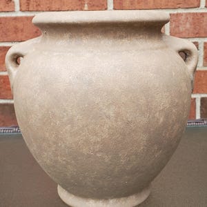 May include: A round, beige-toned ceramic vase with a textured, matte finish. The vessel features a wide, circular opening at the top, a rounded body, and two small, integrated handles on opposite sides, resting on a circular base.