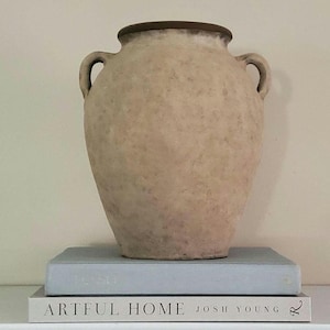 May include: A beige, textured ceramic vase with two side handles and a dark brown rim, resting on a light blue book titled Artful Home by Josh Young. The vase has a mottled, matte finish and a rounded body that tapers toward the base.