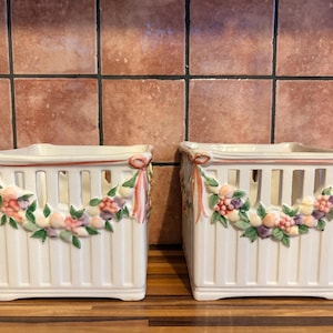 May include: Two square ceramic baskets with a white finish and vertical ribbed sides. Each basket features a decorative relief of colourful fruit garlands and pink ribbons draped across the upper section, with open cut-out slats near the top rim.