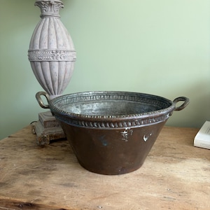 May include: A large, round, hammered copper basin with a wide, flared rim featuring a decorative embossed border. The basin has two small, curved metal handles attached to opposite sides and a weathered, dark brown patina with visible white marks.