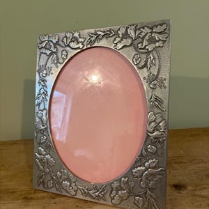 May include: A rectangular silver-toned metal picture frame featuring an embossed floral pattern of poppies and leaves surrounding a central oval opening. The frame has a textured finish and contains a solid light pink insert.