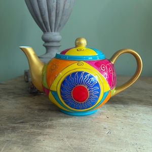 May include: A ceramic teapot featuring a vibrant, multi-coloured geometric pattern with a central blue and red sun motif. The teapot has a gold-painted spout, handle, and lid knob, with additional accents in yellow, pink, orange, and turquoise.