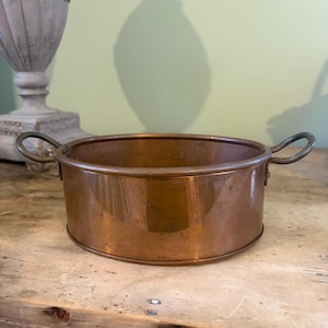 May include: A round copper dish with a polished metallic finish and two curved metal handles attached to opposite sides. The vessel features a smooth, cylindrical wall and a flat base, showcasing a warm, reflective reddish-brown colour throughout.
