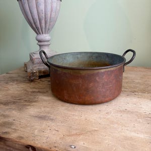 May include: A round, shallow copper pot with a weathered, oxidised patina and two dark metal loop handles on opposite sides. The interior shows signs of age and discolouration, while the exterior displays a textured, brownish-orange metallic surface.