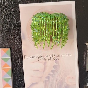 May include: A decorative magnet featuring a three-dimensional potted plant crafted from vibrant green beads. The beaded plant has numerous dangling strands of green beads attached to a small brown base, mounted on a Renue Advanced Cosmetics & Head Spa card.