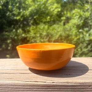 May include: A small, round, smooth ceramic bowl with a vibrant, solid orange glaze. The bowl features a simple, open design with a curved rim and a consistent, glossy finish across its entire exterior surface, reflecting light evenly.