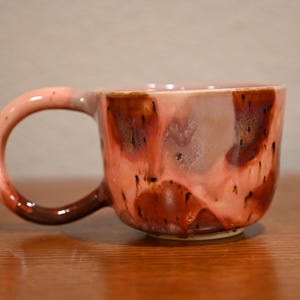 May include: A ceramic mug featuring a rounded body and a curved handle. The exterior surface displays a reactive glaze with blended tones of soft pink, muted lavender, and deep brown, creating a variegated, dripped appearance throughout.