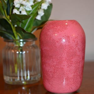 May include: A cylindrical ceramic vase with a rounded top and a vibrant pink glaze featuring a dense, speckled texture of darker red dots across the entire surface. The vase has a smooth, glossy finish and a slightly tapered base.
