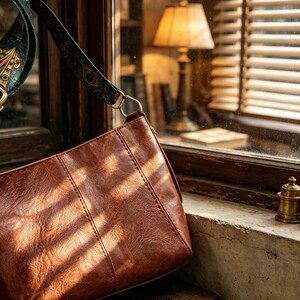 May include: A brown faux leather shoulder bag with a textured surface and vertical stitched detailing. The bag features a detachable fabric strap decorated with a colorful peacock feather pattern in shades of teal, gold, and black, attached with gold-tone metal hardware.