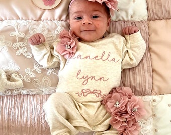 Może przedstawiać: Dziewczynka w beżowym body z wyszytym imieniem "Gianella Lynn". Ma na sobie różową opaskę z kwiatkami i ma różowy kwiatek na body. Dziewczynka leży na różowo-białej patchworkowej kołderce. Na zdjęciu jest też okrągły drewniany znak z napisem "jeden miesiąc".