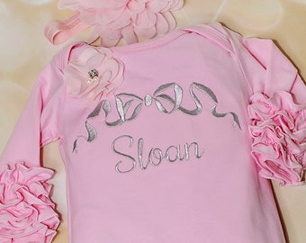 May include: Pink baby onesie with ruffled sleeves and legs. The onesie has a silver embroidered bow and the name "Sloan" on the front. It comes with a pink flower headband.