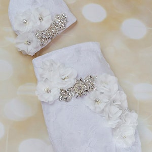 Puede incluir: Un arrullo blanco para bebé con un adorno de flor blanca y strass. El arrullo está hecho de una tela suave y texturizada y tiene un extremo anudado. También se incluye un gorro blanco a juego con un adorno de flor y strass.