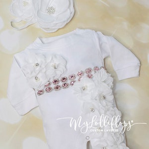 Pode incluir: Macac&atilde;o branco para beb&ecirc; com um cinto de strass rosa e enfeites de flores brancas. O macac&atilde;o tem uma faixa de flores brancas com detalhes de p&eacute;rolas. O texto "My Lolliflops Custom Layette" est&aacute; impresso no macac&atilde;o.