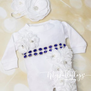 Pode incluir: Um macac&atilde;o branco para beb&ecirc; com um cinto de strass azul e enfeites de flores brancas. O macac&atilde;o tem mangas compridas e bot&otilde;es de press&atilde;o na virilha. Tamb&eacute;m est&aacute; inclu&iacute;do um la&ccedil;o de flores brancas combinando. O texto "My Lollipops Custom Layette" est&aacute; impresso no macac&atilde;o.