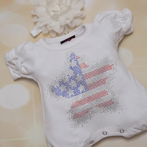 May include: White baby onesie with a red, white and blue star-shaped design made of rhinestones. The star is in the shape of the American flag.