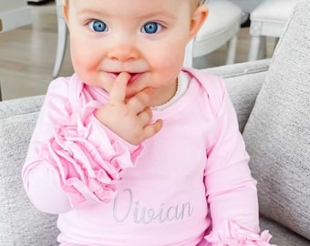 May include: A baby girl wearing a pink ruffled outfit with the name "Vivian" embroidered on the front. She is wearing a pink flower headband and has blue eyes.