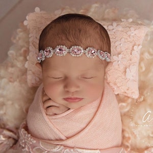 K&ouml;nnte beinhalten: Ein neugeborenes Baby-M&auml;dchen schl&auml;ft friedlich in einem rosa Swaddle, tr&auml;gt ein glitzerndes Stirnband mit rosa Edelsteinen. Das Baby liegt auf einer flauschigen wei&szlig;en und rosa Decke.