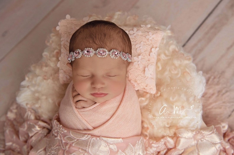 K&ouml;nnte beinhalten: Ein neugeborenes Baby-M&auml;dchen schl&auml;ft friedlich in einem rosa Swaddle, tr&auml;gt ein glitzerndes Stirnband mit rosa Edelsteinen. Das Baby liegt auf einer flauschigen wei&szlig;en und rosa Decke.