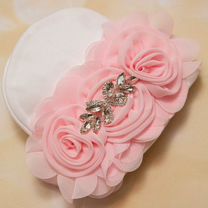 May include: A white fabric headband with three pink fabric roses and a rhinestone embellishment.