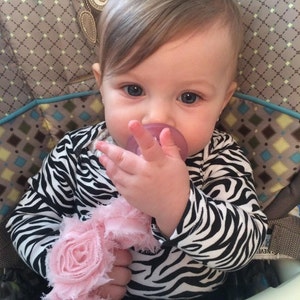 May include: A baby girl wearing a black and white zebra print onesie with a pink flower headband and a pacifier in her mouth. She is sitting in a high chair and has her hand up to her mouth.