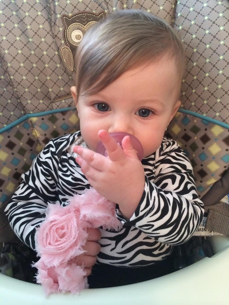 May include: A baby girl wearing a black and white zebra print onesie with a pink flower headband and a pacifier in her mouth. She is sitting in a high chair and has her hand up to her mouth.