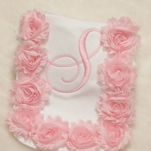 Puede incluir: Un babero blanco para beb&eacute; con una letra "S" bordada en rosa y un ribete de volantes rosa.