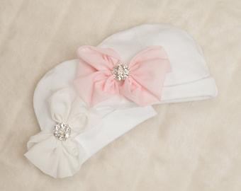 May include: Two white baby hats with bows. One hat has a pink bow with a rhinestone embellishment, and the other has a white bow with a rhinestone embellishment.