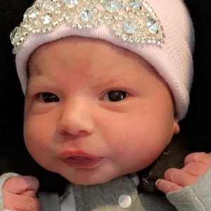 May include: A pink baby hat with a rhinestone design. The hat is on a baby's head.