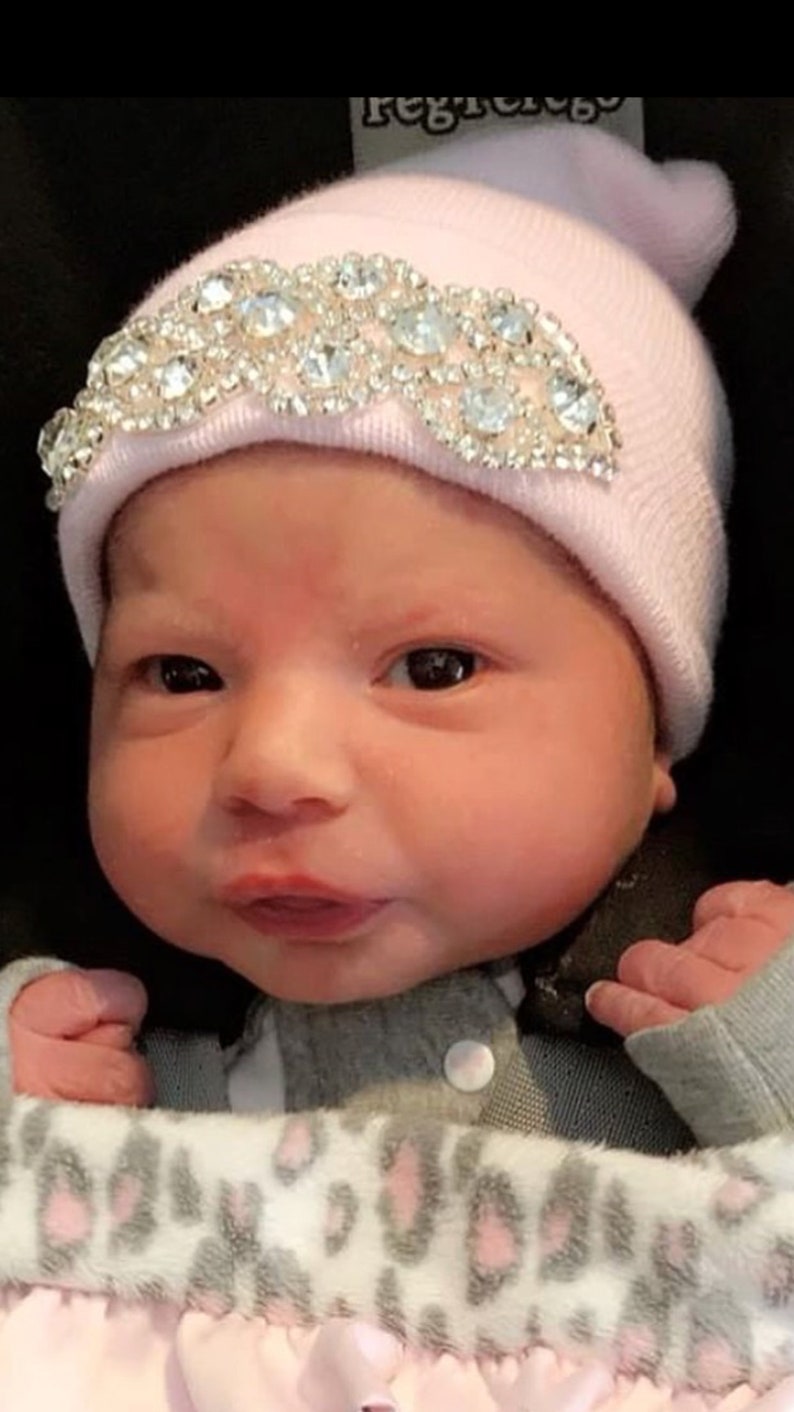 May include: A pink baby hat with a rhinestone design. The hat is on a baby's head.