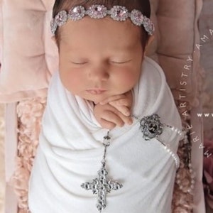 K&ouml;nnte beinhalten: Ein neugeborenes Baby, das in ein wei&szlig;es Swaddle gewickelt ist und ein silbernes und rosafarbenes Strass-Stirnband tr&auml;gt. Das Baby h&auml;lt eine silberne Kreuzkette mit einem Perlen- und Silberanh&auml;nger. Das Baby liegt auf einem rosa und wei&szlig;en Blumenhintergrund.