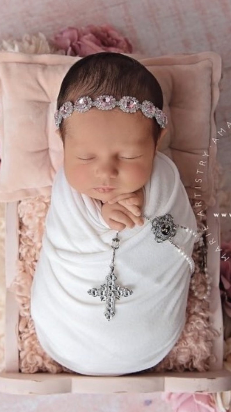 K&ouml;nnte beinhalten: Ein neugeborenes Baby, das in ein wei&szlig;es Swaddle gewickelt ist und ein silbernes und rosafarbenes Strass-Stirnband tr&auml;gt. Das Baby h&auml;lt eine silberne Kreuzkette mit einem Perlen- und Silberanh&auml;nger. Das Baby liegt auf einem rosa und wei&szlig;en Blumenhintergrund.