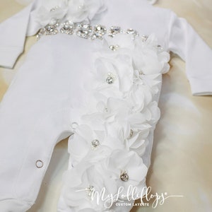 Pode incluir: Macac&atilde;o branco para beb&eacute; com um adorno de flores brancas e um cinto de strass. O macac&atilde;o tem um chap&eacute;u branco a condizer com o texto "Bella Mia" bordado.