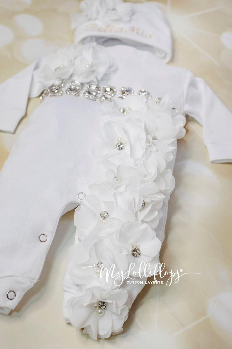 Pode incluir: Macac&atilde;o branco para beb&eacute; com um adorno de flores brancas e um cinto de strass. O macac&atilde;o tem um chap&eacute;u branco a condizer com o texto "Bella Mia" bordado.