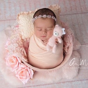 K&ouml;nnte beinhalten: Ein neugeborenes Baby-M&auml;dchen schl&auml;ft in einer rosa Sch&uuml;ssel mit einem rosa Teddyb&auml;r. Das Baby tr&auml;gt ein rosa Stirnband mit Strasssteinen. Die Sch&uuml;ssel ist von rosa Blumen umgeben.