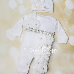 Pode incluir: Macac&atilde;o branco para beb&ecirc; com cinto de strass e enfeites de flores brancas. O macac&atilde;o tem o texto "My Lollipops" na perna. Um chap&eacute;u branco com um enfeite de flor e o texto "Bella Mia" est&aacute; inclu&iacute;do.