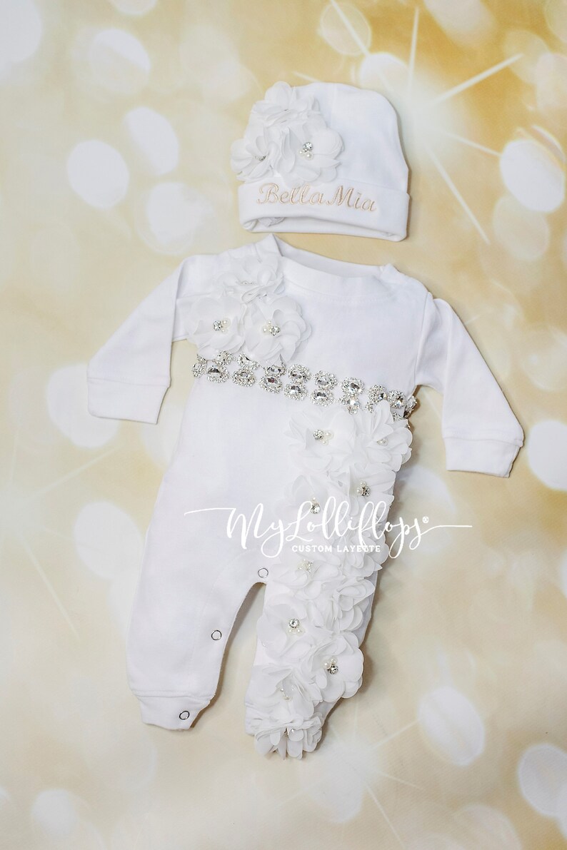 Pode incluir: Macac&atilde;o branco para beb&ecirc; com cinto de strass e enfeites de flores brancas. O macac&atilde;o tem o texto "My Lollipops" na perna. Um chap&eacute;u branco com um enfeite de flor e o texto "Bella Mia" est&aacute; inclu&iacute;do.