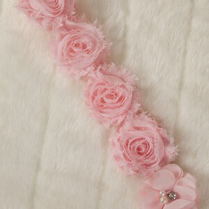 May include: A row of five pink fabric flowers with ruffled edges, arranged in a line on a white fuzzy surface. The last flower has a small pearl and rhinestone embellishment in the center.