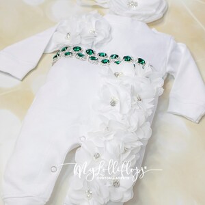 Pode incluir: Macac&atilde;o branco para beb&ecirc; com um cinto de strass verde e enfeites de flores brancas. O macac&atilde;o tem uma faixa combinando com flores brancas.