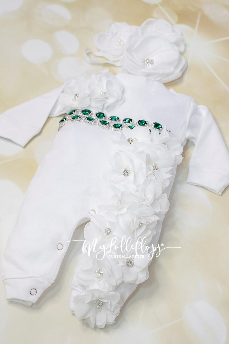 Pode incluir: Macac&atilde;o branco para beb&ecirc; com um cinto de strass verde e enfeites de flores brancas. O macac&atilde;o tem uma faixa combinando com flores brancas.