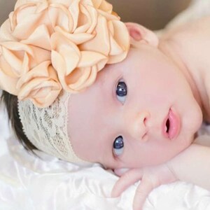 以下が含まれることがあります： 白い布の上に横たわっている、桃色の花柄のレースのついたヘッドバンドをつけた女の子の赤ちゃん。赤ちゃんは青い目をしています。